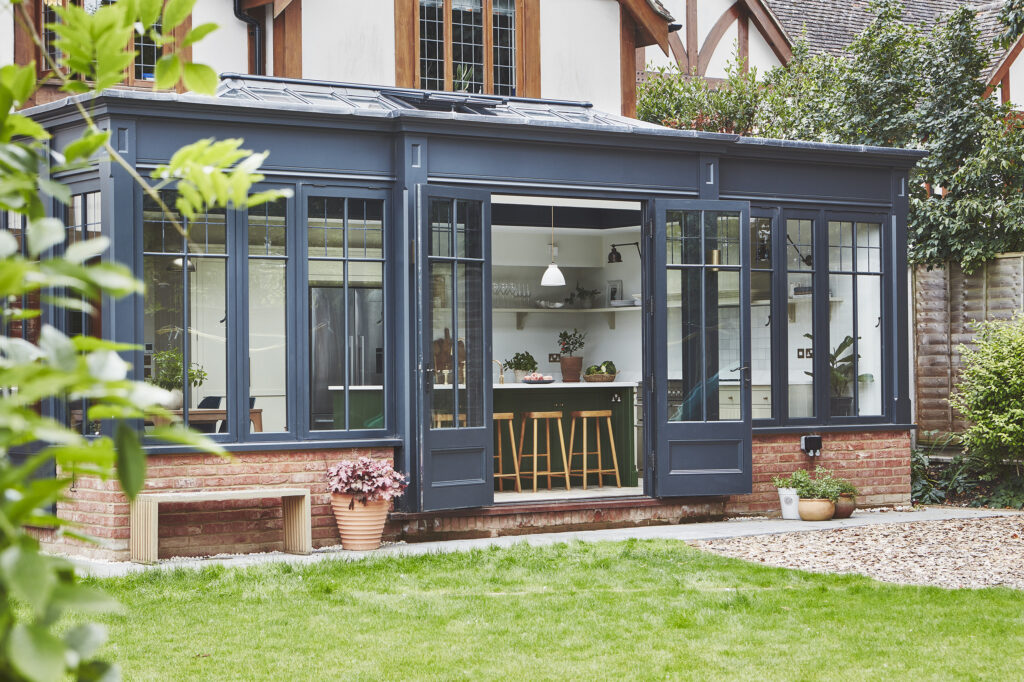 A stylish kitchen with a green island set within a modern ornate orangery overlooking a lush garden.