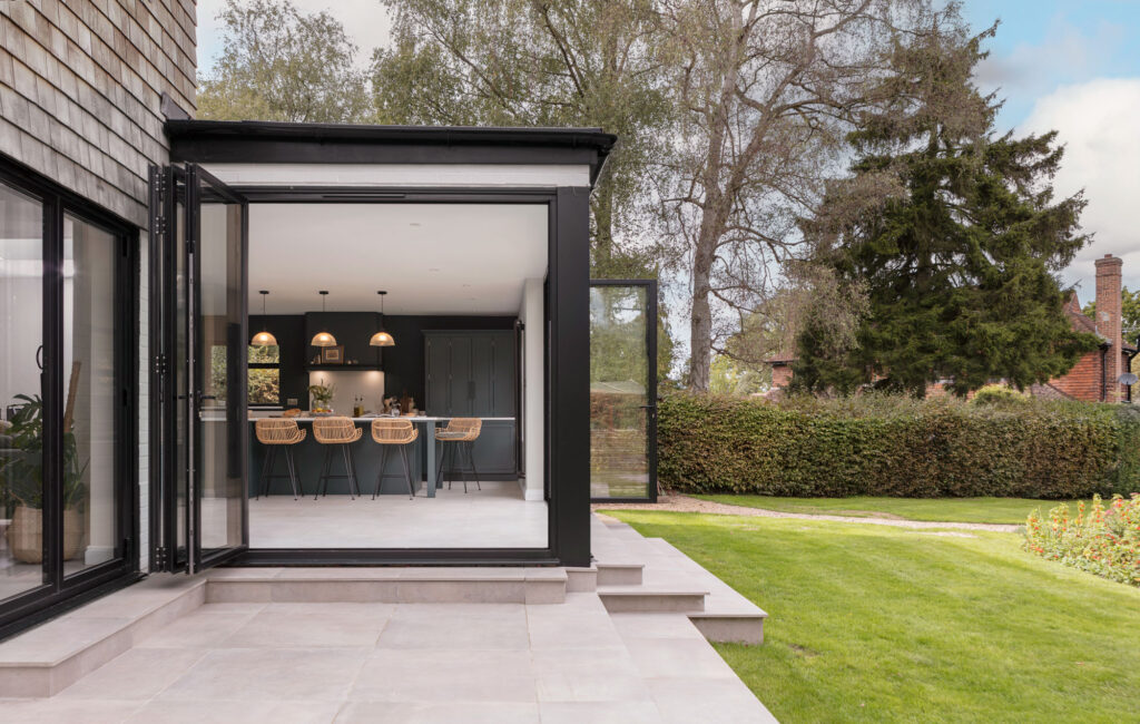 Modern house extension with open patio doors, leading to a beautiful green kitchen with bar stools.