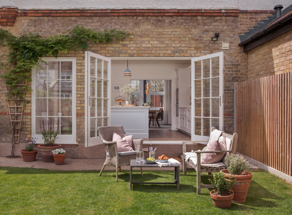 Cosy home patio with wicker chairs, and potted plants. Open French doors leads to a stylish white kitchen.