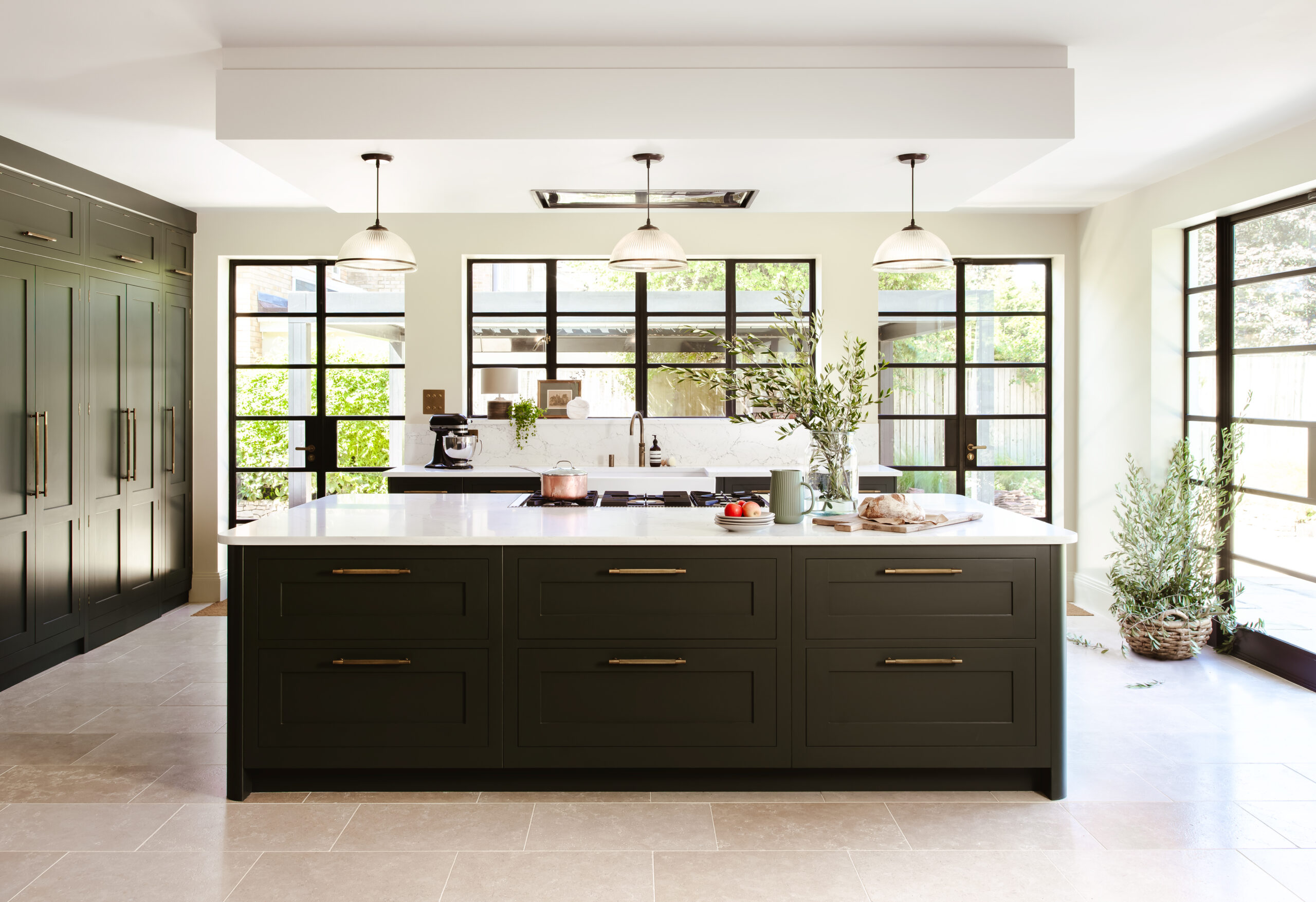 Green kitchen unit and worktop with feruit on top in front of 3 windows